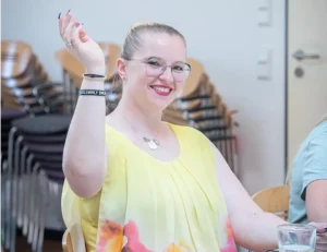 Eine freundliche Mitarbeiterin vom Pflegedienst Kremer lächelt in die Kamera und hebt die Hand, während sie hochwertige Pflege im Team repräsentiert.