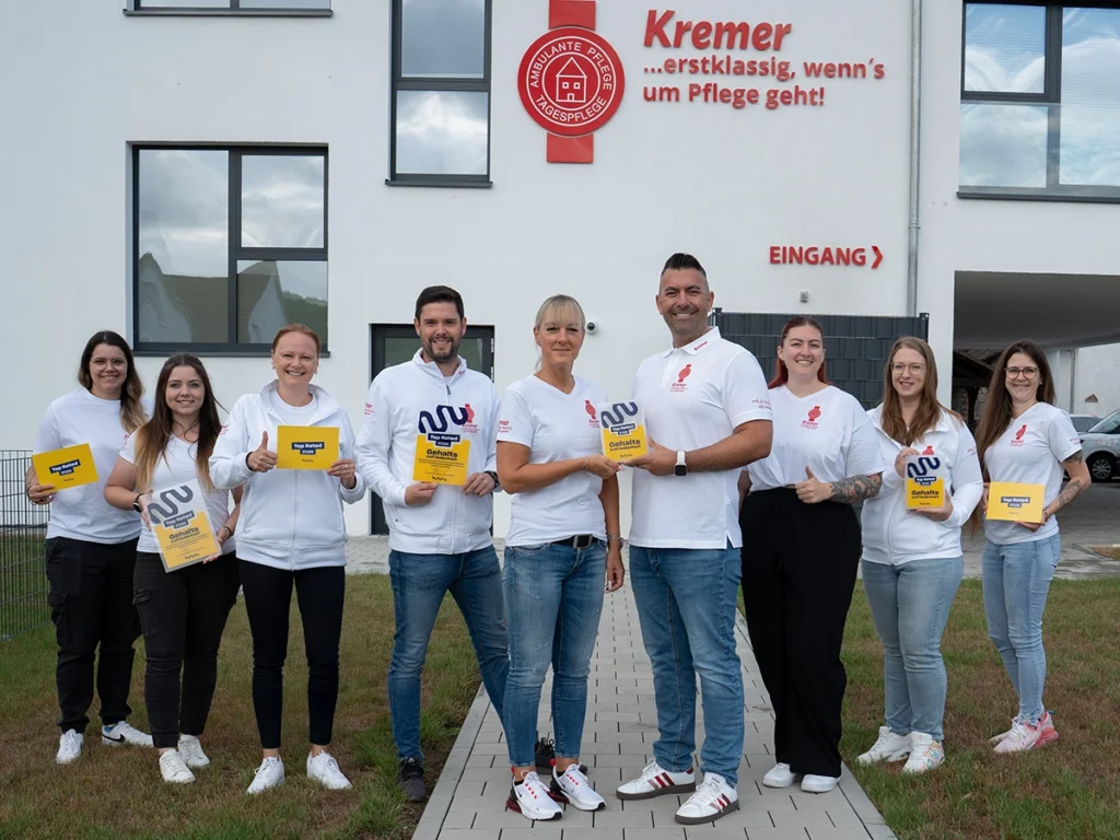 Neun Personen vom Team stehen nebeneinander in einer Reihe draußen vor dem Kremer Gebäude und halten jeweils eine Siegelbenachrichtigung in der Hand. Dabei lächeln sie in die Kamera.