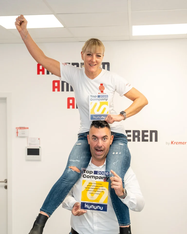 Mitarbeiterin auf der Schulter vom Chef. Beide in Siegerpose und jeweils ein Auszeichnungsiegel in der Hand.