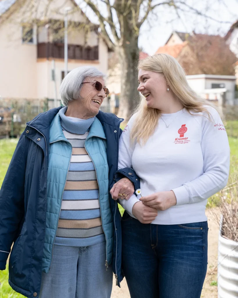 Eine freundliche Pflegekraft vom Pflegedienst Kremer begleitet eine ältere Dame bei einem Spaziergang im Freien für die ambulante Pflege MKK.