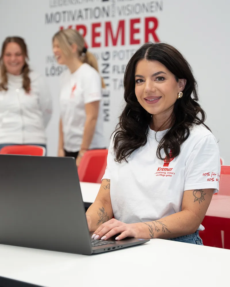 Eine Mitarbeiterin vom Pflegedienst Kremer arbeitet am Laptop in einem Büro, während im Hintergrund weitere Kollegen für die Pflegeberatung lächeln.