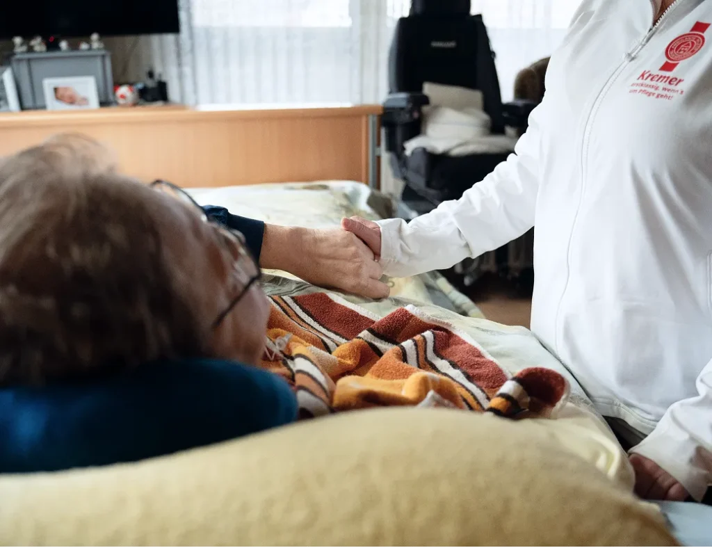 Eine Pflegekraft vom Pflegedienst Kremer hält die Hand eines Patienten im Bett bei der ambulanten Pflege im Main-Kinzig-Kreis für mehr Geborgenheit.