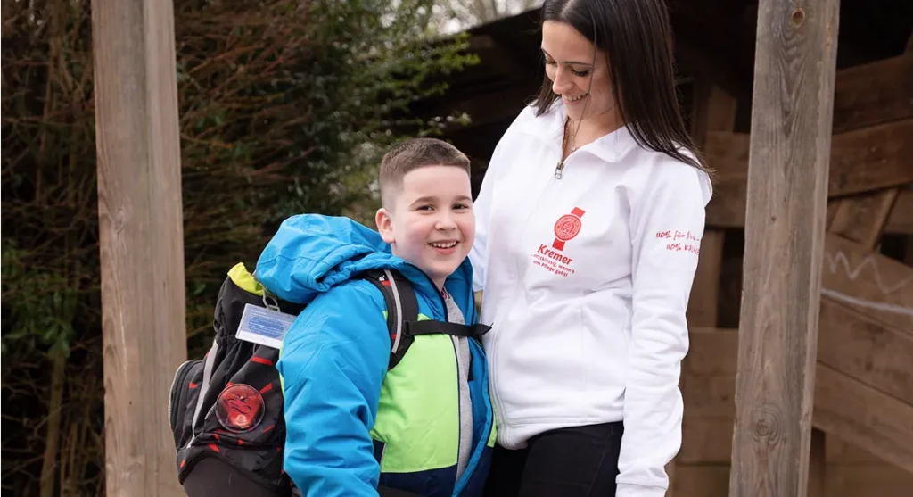 Eine Pflegekraft vom Pflegedienst Kremer begleitet ein glückliches Kind mit Schulranzen im Rahmen der hochwertigen Kinderintensivpflege im Freien.