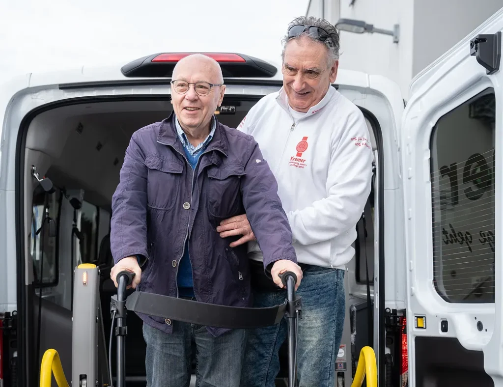 Ein Mitarbeiter vom Pflegedienst Kremer unterstützt einen älteren Klienten mit Rollator beim Aussteigen aus einem barrierefreien Fahrzeug.