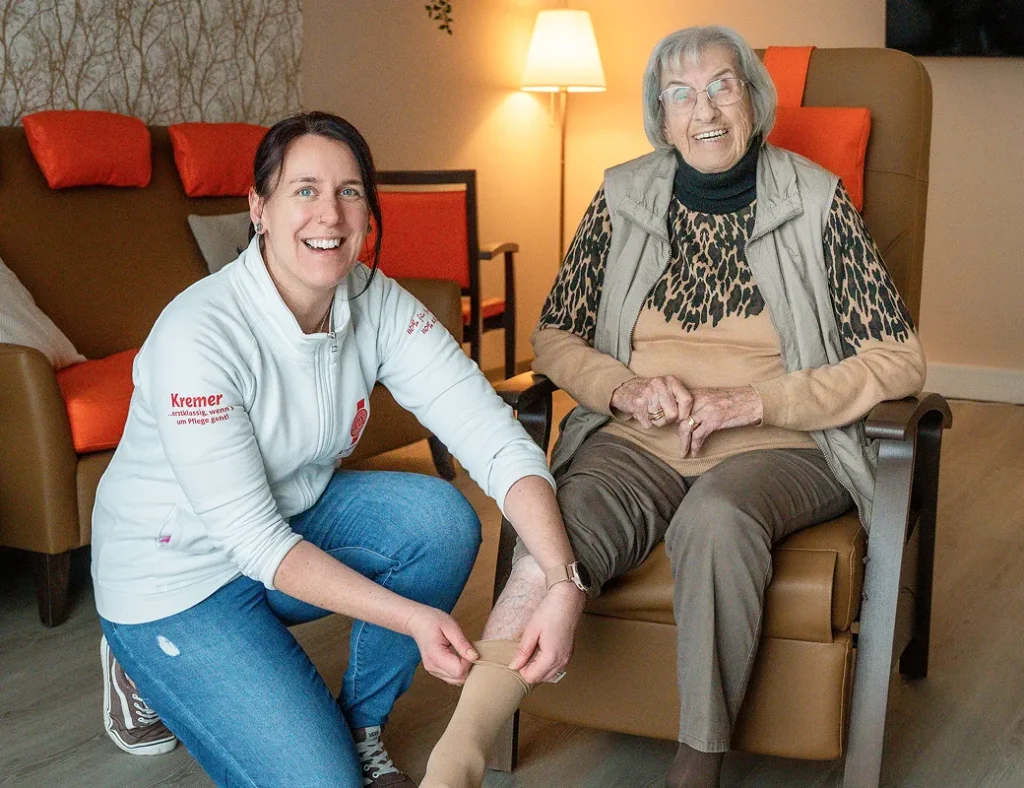 Eine freundliche Pflegekraft vom Pflegedienst Kremer unterstützt eine Seniorin bei der ambulanten Pflege im MKK durch das Anlegen eines Kompressionsstrumpfes.