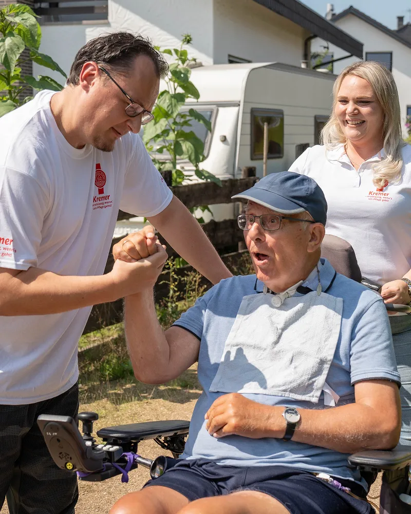 Pflegekraft von Pflegedienst Kremer unterstützt einen älteren Klienten im Rollstuhl bei der außerklinischen Intensivpflege im Freien mit viel Freude.