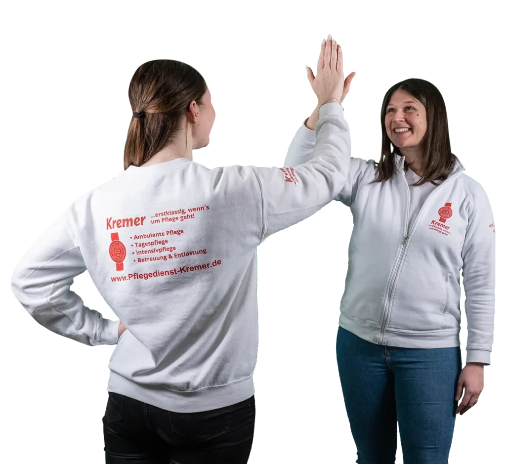 Zwei Mitarbeiterinnen vom Pflegedienst Kremer im Main-Kinzig-Kreis geben sich ein High-Five und präsentieren ihre Arbeitskleidung mit Firmenlogo.