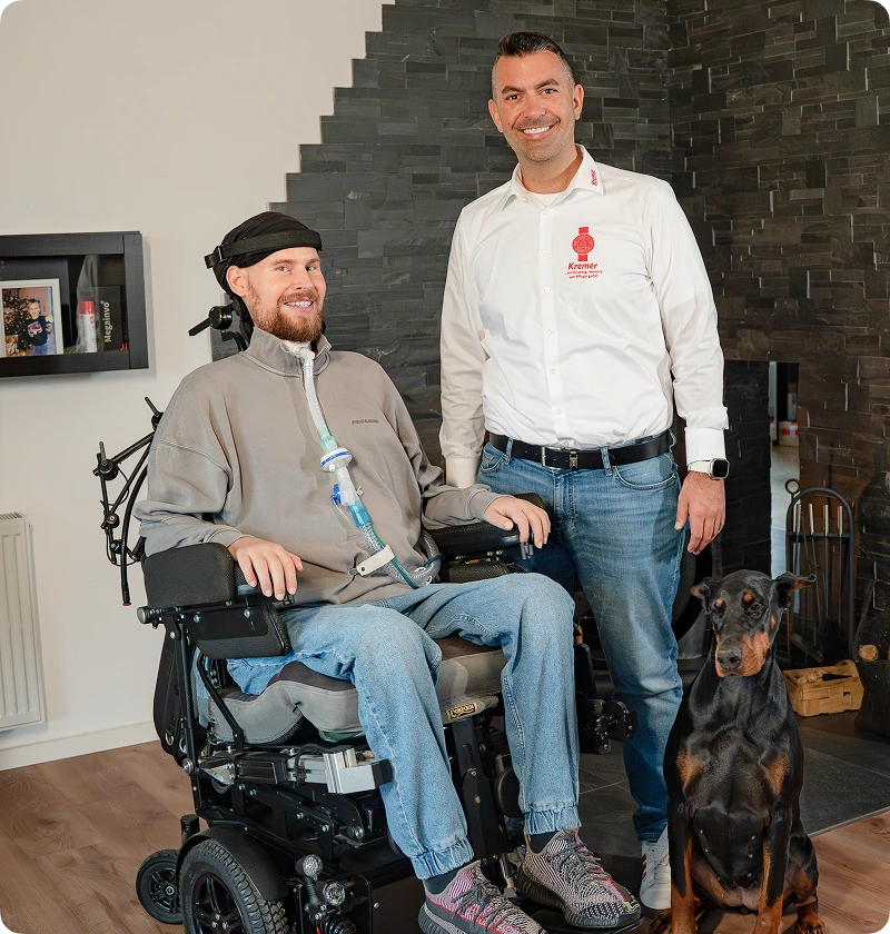 Patrick Kremer mit Hund und mit einem Klienten im Rollstuhl neben ihm in einem Treppenhaus.