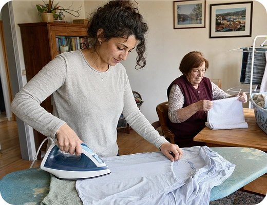 Eine Pflegekraft vom Pflegedienst Kremer unterstützt eine Seniorin bei der Hausarbeit durch Bügeln für eine hochwertige Pflege im häuslichen Umfeld.